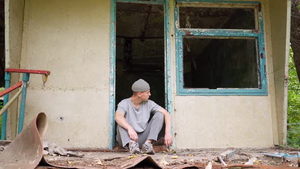 A sad homeless guy sits on the porch of an abandoned house. Bad neighborhood and poverty alt