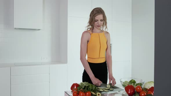 Young Woman Slicing Cucumber For Vegetable Salad. White Modern Kitchen.