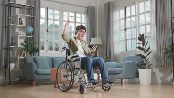 Asian Man Sitting In A Wheelchair While Use Mobile Phone Playing Video Game And Celebrating At Home alt