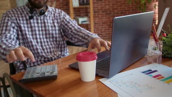 Disabled Person in Wheelchair is Typing on Laptop and Counting on Calculator There is Cup of Coffee alt