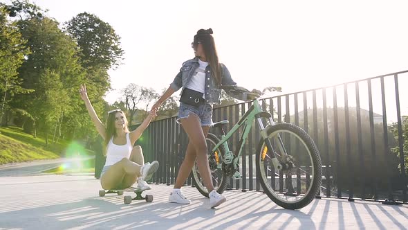 Young Woman Sitting on a Longboard while Her Best Female Friend Goes Ahead of Her alt