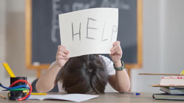 Exhausted Cute Schoolboy Putting Head on Desk and Raising Help Written on Paper alt