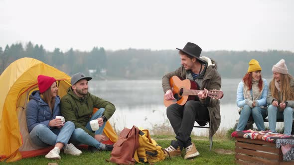Group of Friends camping.They Are Sitting Around Camp Fire, Playing Guitar alt