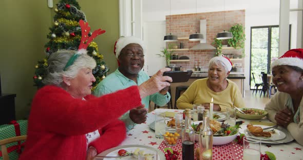 Diverse senior friends in santa hats looking at smartphone and laughing at christmas dinner table alt