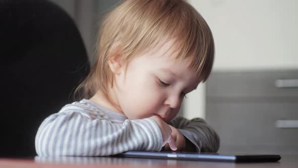 A Small Child of 23 Years Old Sitting on a Chair at the Table Looking at the Tablet and Tapping 