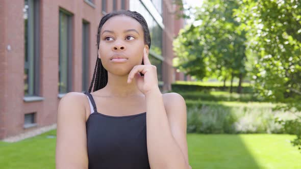 A Young Black Woman Thinks About Something - an Office Building and Green Park in the Background alt