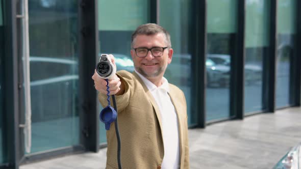 Older Man Stands with Charger Near His Electric Car alt