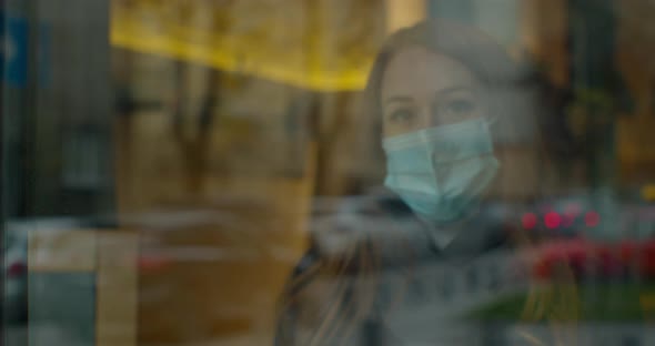 Young Woman Wears a Mask To Protect Herself From Covid-19 While Looking Out the Window. alt