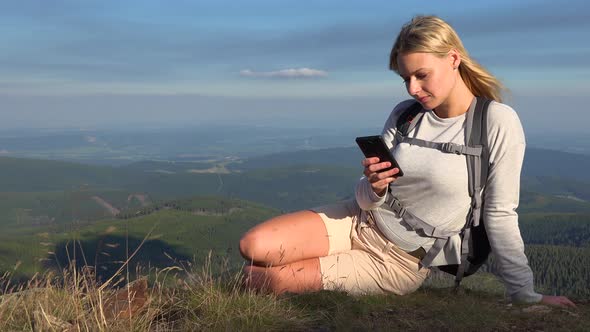 A Young Beautiful Woman Sits on Grass on a Hilltop and Works on a Smartphone alt
