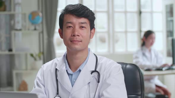 Young Asian Man Doctor Is Using Laptop Computer, He Turns And Smiles To Camera In Workplace alt