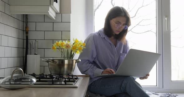 Hipster female in eye glasses working on computer from home kitchen. Multitasking concept.   alt