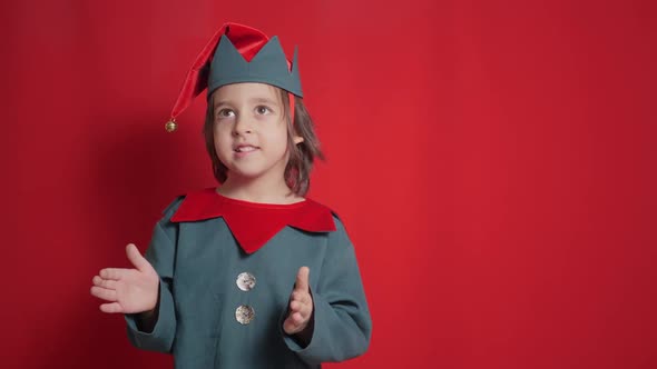 Boy Child in Dwarf Costume Stands on a Red Background Catching a Gift Box alt
