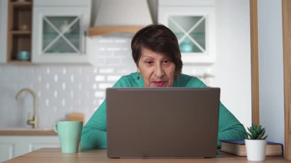 Happy Elderly Woman Communicating By Video Call Using Laptop at Home in Kitchen alt