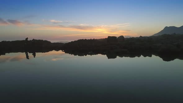 Drone slowly flies low over natural ocean pool, rising to reveal the stunning sunset behind the moun alt