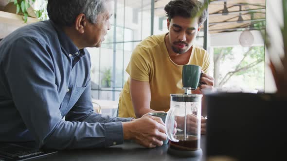 Man talking to young son drinking coffee from mug while leaning on kitchen counter at home alt