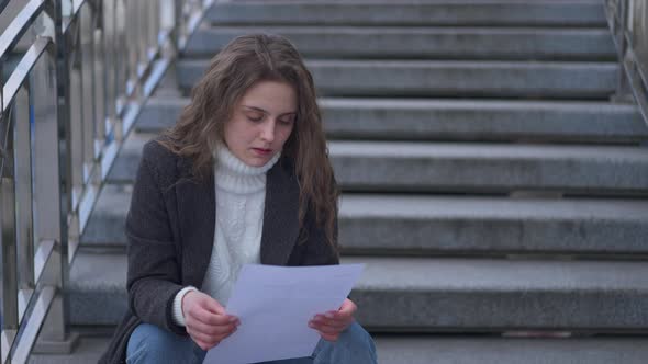 Young Brunette Caucasian Sad Woman Reading Letter Tearing Paper Sitting on Urban City Stairs alt