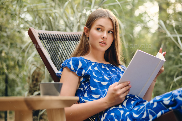 Young beautiful woman in blue dress dreamily reading book on wooden deck chair in park