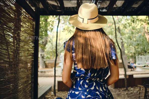 Young pretty brown haired woman in blue dress and hat from back dreamily standing in beautiful park