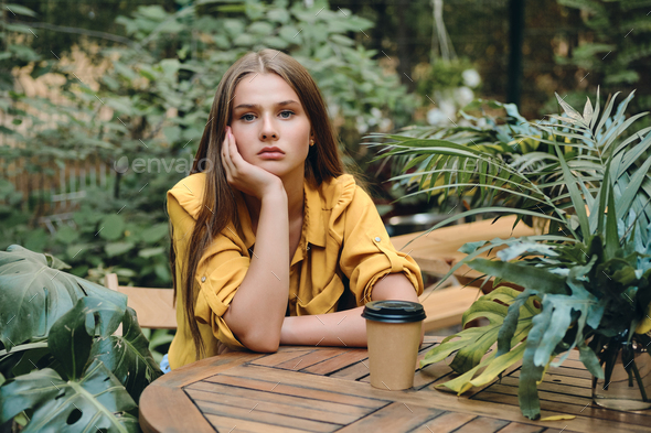 Girl in yellow shirt sadly looking in camera sitting with cup of coffee to go in beautiful city park