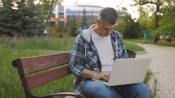 A Male Freelancer Sits on a Bench in a Park and Works on a Laptop