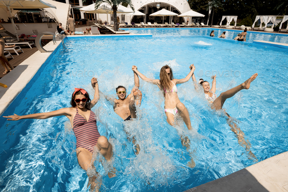 Company of young merry girls and guy jump together holding hands in the swimming pool next to lounge