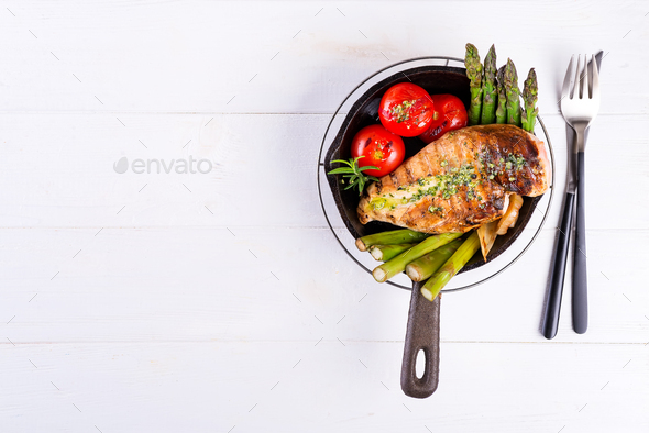 Grilled chicken breast on a cast iron skillet with grill vegetables on a wooden background, flat lay