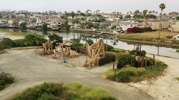An aerial view rotates around a set of oil pumpjacks along a beachfront channel with houses nearby. alt