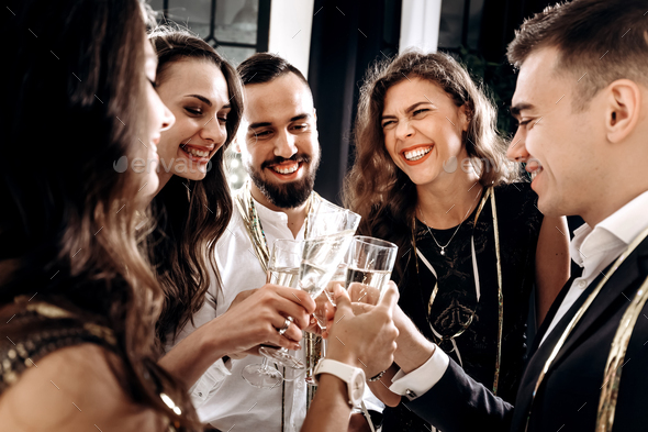 Company of friends dressed in stylish elegant clothes stand together and clink glasses with