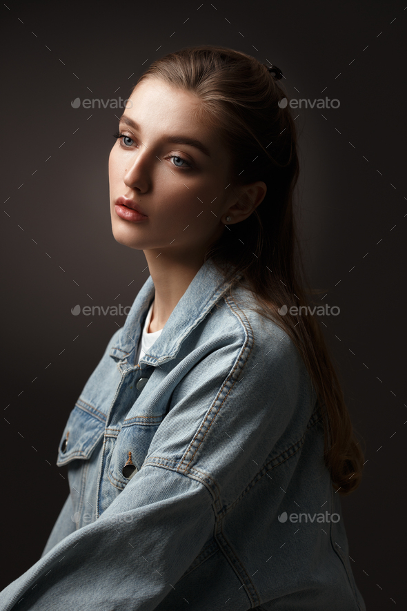 Portrait of beautiful brunette girl with hair tied back dressed in jeans jacket on the dark