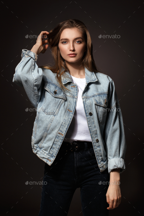 Beautiful brunette girl with long flowing hair dressed in jeans jacket and jeans  poses holding her hands on her head on the dark background in the studio  Stock Photo  Adobe Stock