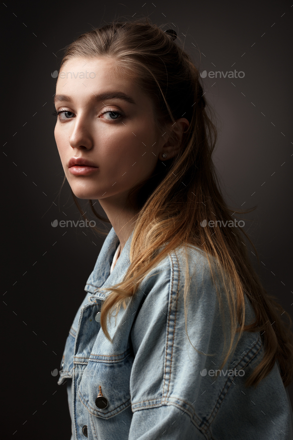 Portrait of beautiful brunette girl with hair tied back dressed in jeans jacket on the dark