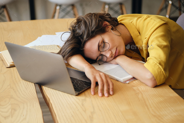 Tired woman in eyeglasses fall asleep on desk with laptop and documents under head at workplace