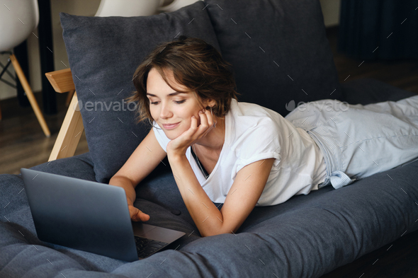 Young beautiful smiling woman lying on sofa dreamily working on laptop in modern co-working space