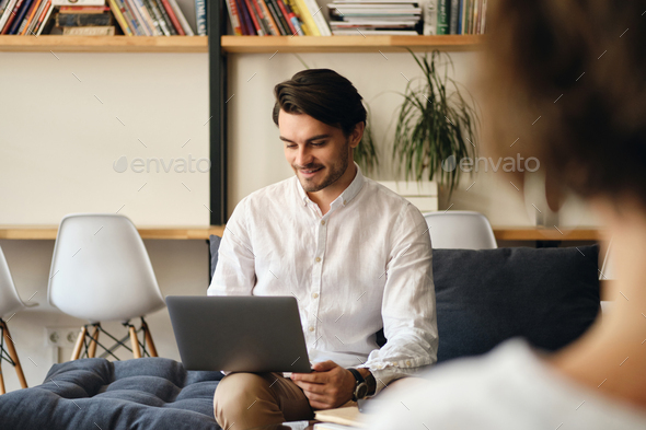 Young smiling businessman on sofa with colleague happily working on laptop in co-working space