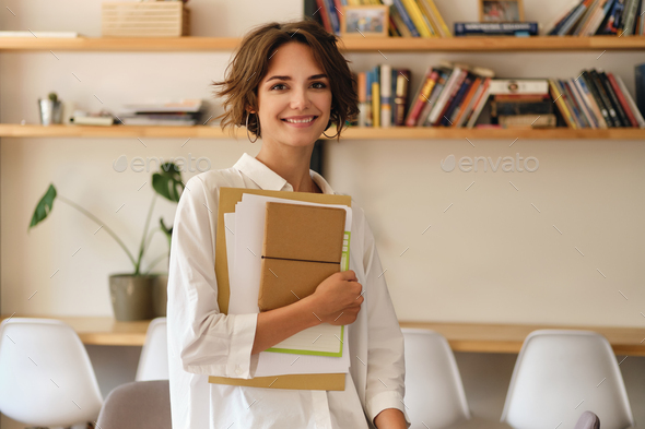 Young attractive smiling woman joyfully looking in camera with papers and notepad in office