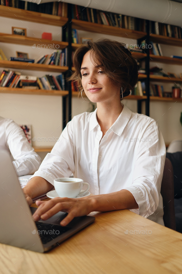 Beautiful smiling woman sitting at the table happily working on laptop with cup of coffee in office