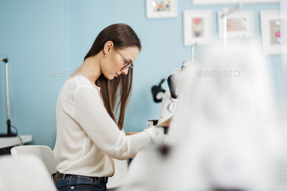 A smart-looking pretty brunet woman wearing white shirt is sewing with the electric sewing-machine - Stock Photo - Images