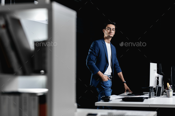 Stylish professional architect in glasses dressed in a blue checkered jacket stands next to the desk