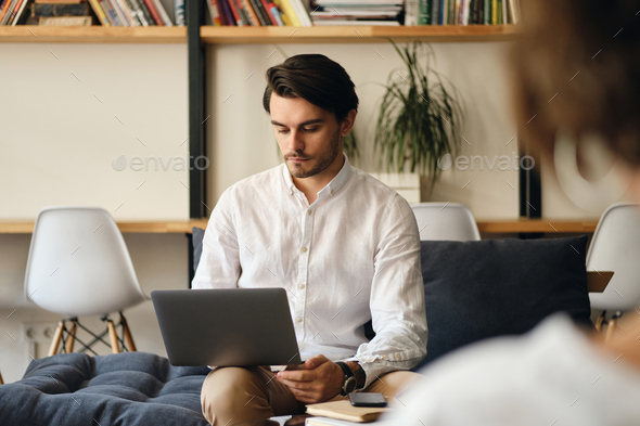 Handsome businessman sitting on sofa thoughtfully working on laptop in modern co-working space