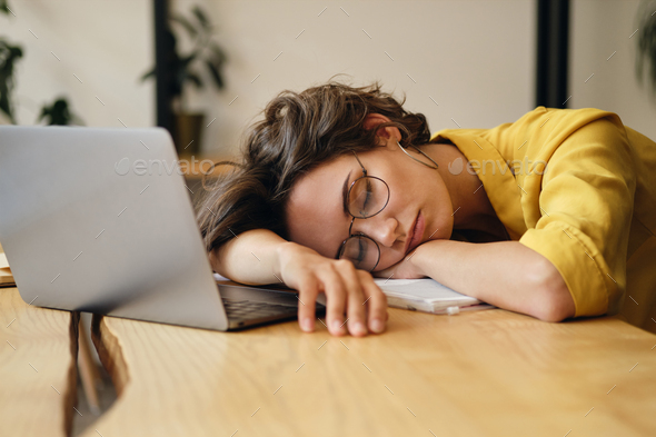 Young tired woman in eyeglasses sleeping on desk with laptop at workplace