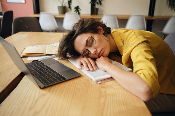 Young tired woman in eyeglasses fall asleep on desk with laptop and notepad under head at workplace