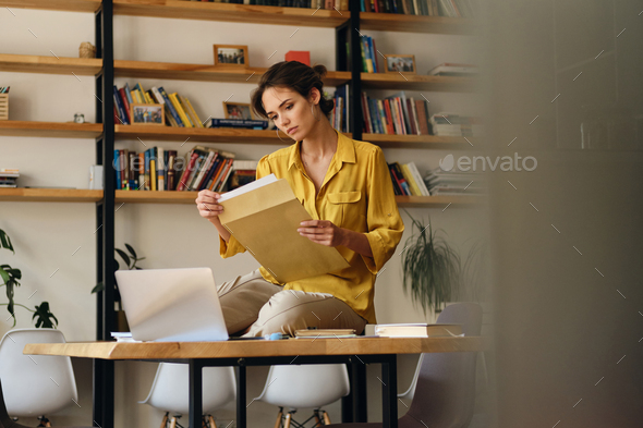 Beautiful woman in shirt sitting on desk with documents thoughtfully working in modern office - Stock Photo - Images