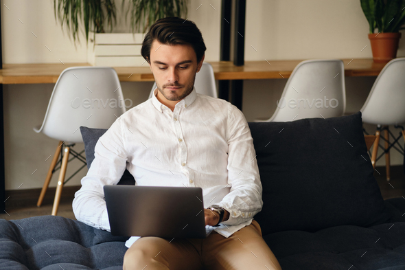 Attractive businessman sitting on sofa thoughtfully working on laptop in modern co-working space