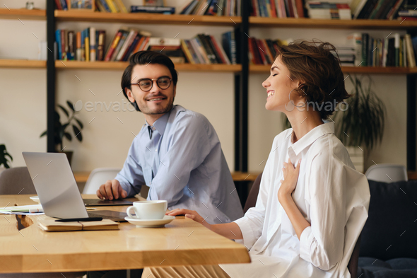 Young business colleagues sitting at the table with laptop happily talking at work in office