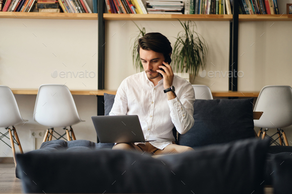 Handsome businessman sitting on sofa thoughtfully talking on cellphone working on laptop - Stock Photo - Images