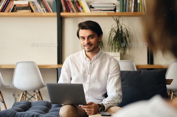 Attractive smiling businessman sitting on sofa with laptop happily looking in camera in co-working