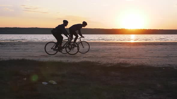 Two Young Men Riding Bicycles on the Beach on the Background of an Orange Sunsetting Sky Slow Motion alt