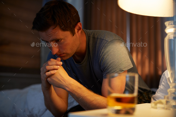Depressed Man Wearing Pajamas Sitting On Side Of Bed With Glass Of Whisky On Bedside Cabinet