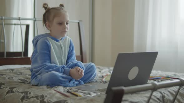 Little Schoolgirl Uses Computer Sitting on the Bed at Home