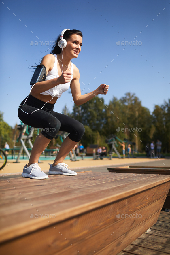 Cheerful woman in headphones doing jump squat on step